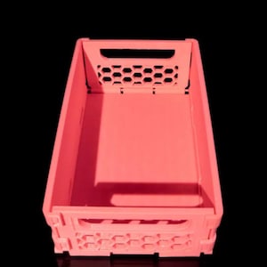 Puede incluir: Una caja de plástico de color coral con forma rectangular. La caja presenta un patrón de panal en los laterales y la parte superior, con una abertura para el asa. El fondo es negro.