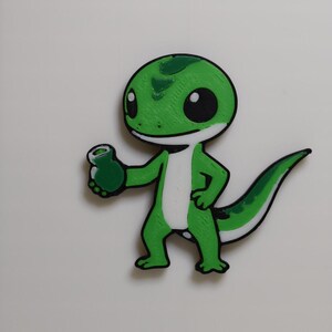 May include: A green cartoon gecko holding a small green object. The gecko has a white belly, black eyes, and a green and white tail. The gecko is outlined in black and is on a white background.
