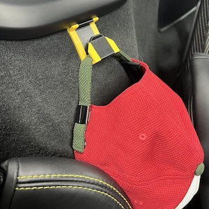 May include: A red baseball cap with a white brim is held by a yellow and black clip attached to a car seat. The clip has a green strap. The car seat is black with yellow stitching.