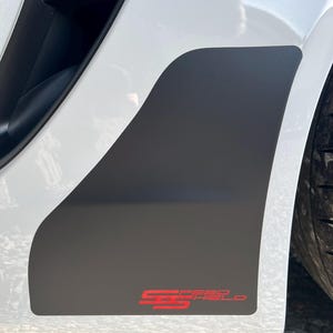 Könnte beinhalten: Nahaufnahme eines weißen Autos mit einem schwarzen Akzentpaneel. Auf dem Paneel steht in roter Schrift "SPEED SHIELD" in einem stilisierten Schriftzug. Das Rad des Autos ist teilweise sichtbar.