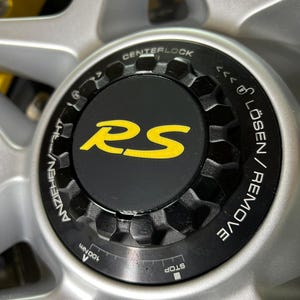 May include: Close-up of a silver car wheel with a black center cap. The center cap has the yellow letters "RS" and the word "CENTERLOCK" is visible. The wheel has a hexagonal pattern around the center cap.