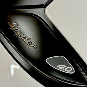 Könnte beinhalten: Nahaufnahme eines schwarz-weißen Autodetails. Das Wort "Spyder" ist in goldener Schrift geschrieben. Ein schwarzes Emblem mit "4.0" in Weiß ist ebenfalls sichtbar. Die Karosserie des Autos ist weiß.