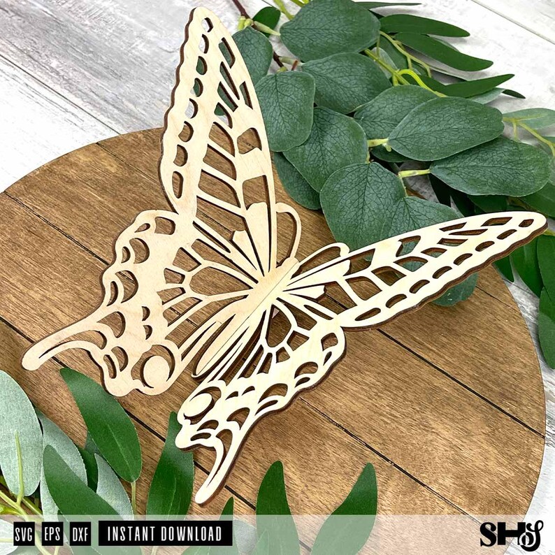 May include: A laser-cut wooden butterfly cutout featuring intricate, open-work wing patterns. The light-colored wood butterfly is positioned on a circular wooden base, with green eucalyptus leaves visible around the edges of the cutout.