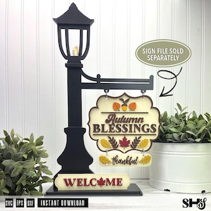 Op de afbeelding: Een zwarte decoratieve lantaarnpaalstandaard met een hangend bord met de tekst Autumn BLESSINGS en thankful, en een basisbord met de tekst WELCOME en een esdoornblad. Het bord bevat herfstblad- en eikelaccenten in oranje en bruin.