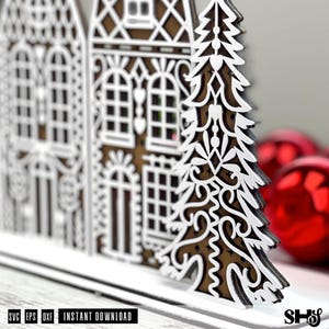 May include: A layered laser-cut decoration featuring a row of two houses and a pine tree with intricate white filigree patterns over a brown base. The text SVG EPS DXF INSTANT DOWNLOAD is visible at the bottom left, with SHS at the bottom right.