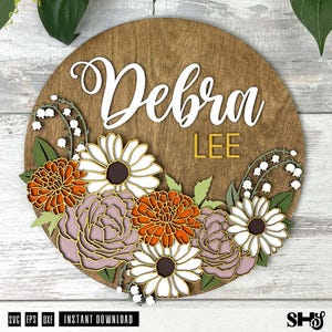 K&ouml;nnte beinhalten: Ein rundes Holzschild mit dem Namen "Debra Lee" in wei&szlig;en Buchstaben auf braunem Hintergrund. Das Schild ist mit einem Blumenkranz aus wei&szlig;en, orangefarbenen und rosa Bl&uuml;ten verziert.