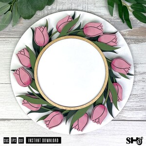 May include: A circular wooden sign featuring a layered design of pink tulips and dark green leaves arranged in a wreath pattern around a central blank white circle. The sign has a natural light wood border and a smooth white background finish.