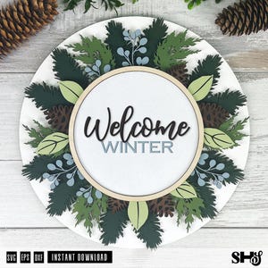 May include: A circular wooden sign featuring the text Welcome WINTER in black and light blue script and block lettering. The sign is decorated with layered cutouts of dark green pine branches, light green leaves, brown pinecones, and light blue berries.