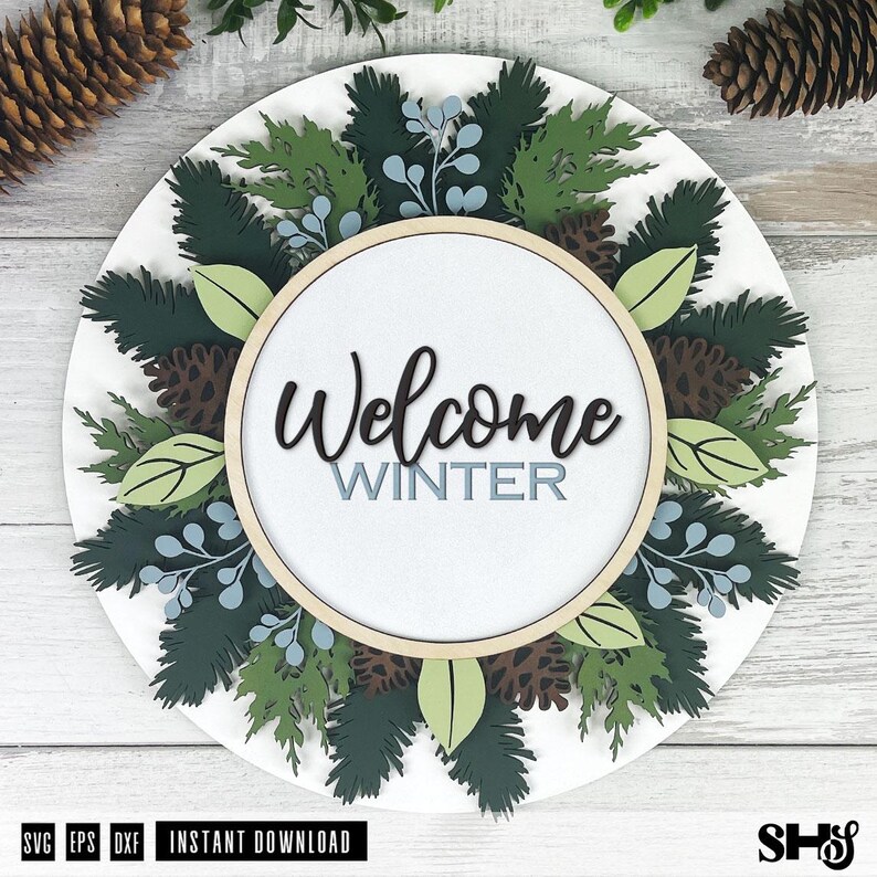 May include: A circular wooden sign featuring the text Welcome WINTER in black and light blue script and block lettering. The sign is decorated with layered cutouts of dark green pine branches, light green leaves, brown pinecones, and light blue berries.
