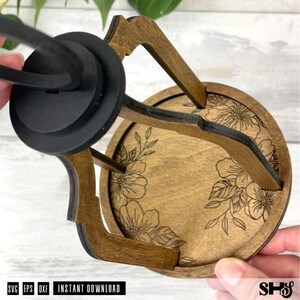 May include: A circular wooden tray with a dark brown finish featuring a laser-engraved floral pattern on the base. The tray has a black circular handle attached to a frame of four wooden supports extending upward from the tray's edge.