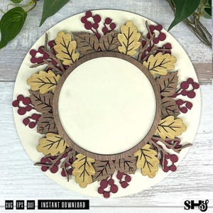 May include: A circular wooden sign featuring a layered design with a central blank circle surrounded by gold and dark brown oak leaves and clusters of maroon berries. The sign is mounted on a light cream circular base with text at the bottom.