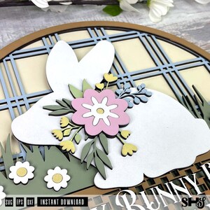 Pode incluir: Uma placa de parede de madeira em camadas com a silhueta de um coelho branco sobre um fundo xadrez azul claro e creme. O coelho &eacute; decorado com uma flor rosa, folhas verdes e pequenas flores amarelas, com flores brancas na grama verde.