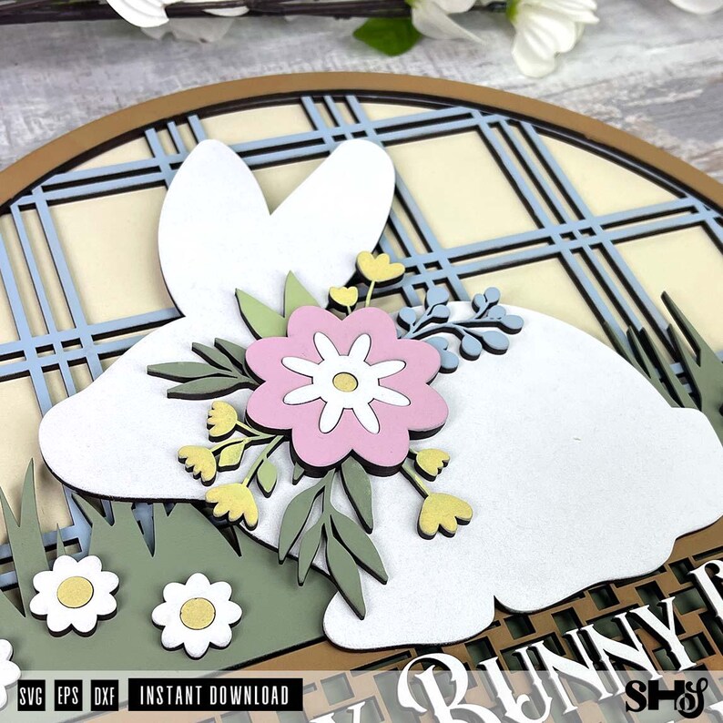 Pode incluir: Uma placa de parede de madeira em camadas com a silhueta de um coelho branco sobre um fundo xadrez azul claro e creme. O coelho &eacute; decorado com uma flor rosa, folhas verdes e pequenas flores amarelas, com flores brancas na grama verde.