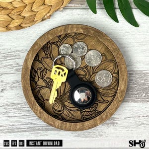May include: A round wooden tray with a dark brown finish and an engraved floral pattern on the base. Inside the tray are four silver-colored coins, a bright yellow key, and a black circular tracking device attached to a silver metal key ring.