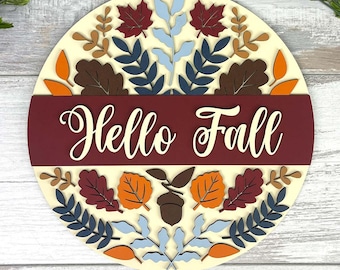 May include: A round wooden sign with the words "Hello Fall" in white script. The sign is cream-colored with a burgundy banner. It is decorated with fall leaves in shades of red, orange, brown, and blue. The sign is a seasonal decoration.