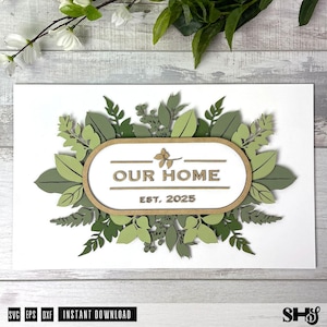 May include: A white sign with a brown wooden frame and green leafy foliage around the edges. The sign says "Our Home Est. 2025".