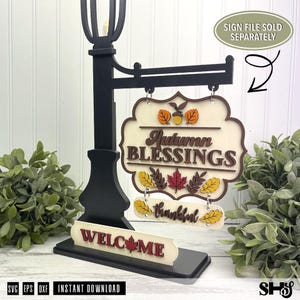 Op de afbeelding: Een zwarte standaard in de stijl van een lantaarnpaal met een hangend bord met de tekst Autumn BLESSINGS en een kleiner bord eronder met het woord thankful. De voet bevat een WELCOME-bord met een esdoornblad.