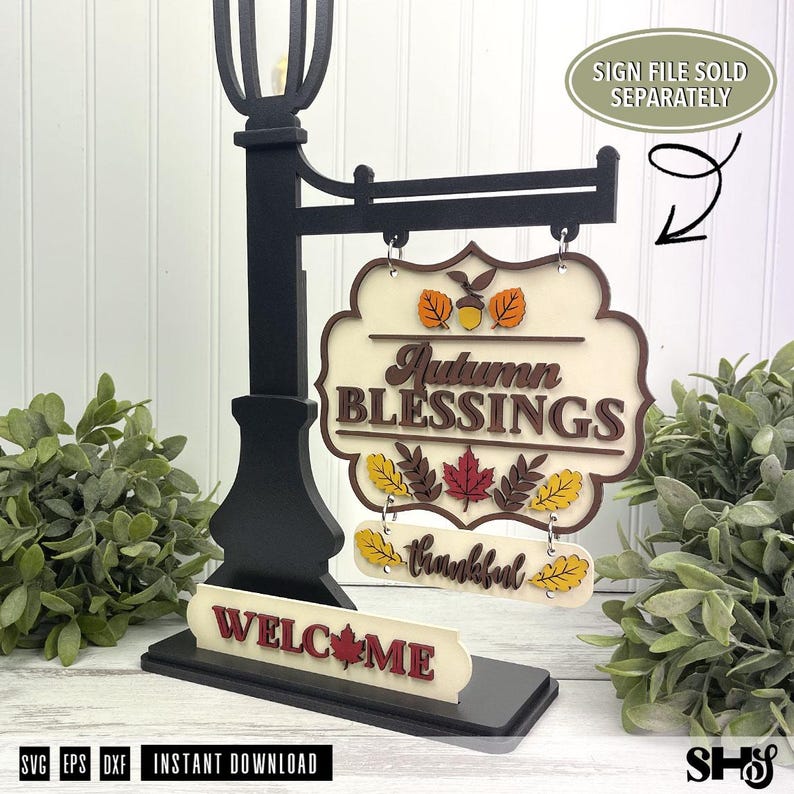 Op de afbeelding: Een zwarte standaard in de stijl van een lantaarnpaal met een hangend bord met de tekst Autumn BLESSINGS en een kleiner bord eronder met het woord thankful. De voet bevat een WELCOME-bord met een esdoornblad.