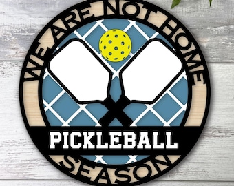 May include: A black and white round sign with the text "We are not home Pickleball Season" in a white font. The sign has a blue and white checkered pattern with two black pickleball paddles and a yellow pickleball in the center.