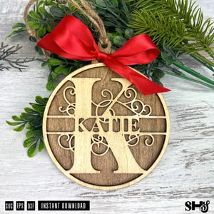 Op de afbeelding: Een ronde houten ornament met een grote uitgesneden letter K met de naam Katie in het midden verwerkt. Het ontwerp bevat decoratieve krullen en een felrood satijnen lint dat aan de bovenkant tot een strik is gebonden.