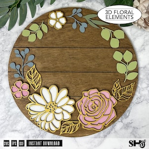May include: A circular wooden sign featuring a dark brown horizontal plank design decorated with layered 3D floral elements. The arrangement includes white and pink flowers, gold-toned filigree leaves, and light green and blue foliage accents.