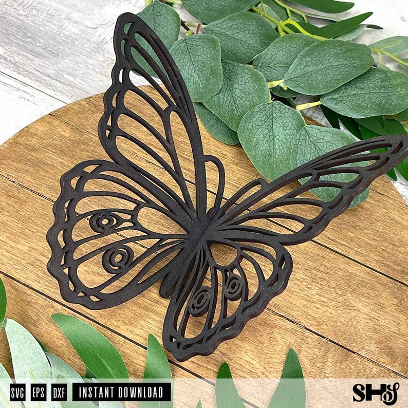 May include: A dark brown, flat, laser-cut butterfly silhouette featuring intricate, open-work wing patterns with circular details. The butterfly is positioned on a circular wooden surface, with green eucalyptus leaves visible around the edges.