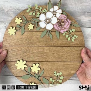 Puede incluir: Un letrero circular de madera con detalles de tablones horizontales. El letrero est&aacute; decorado con elementos florales de madera cortados con l&aacute;ser en capas, en tonos blanco, rosa suave y amarillo p&aacute;lido, con hojas y tallos de color verde salvia.