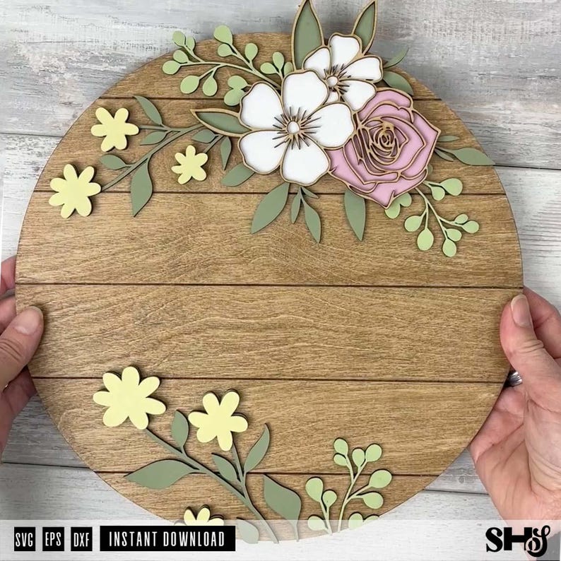 Puede incluir: Un letrero circular de madera con detalles de tablones horizontales. El letrero est&aacute; decorado con elementos florales de madera cortados con l&aacute;ser en capas, en tonos blanco, rosa suave y amarillo p&aacute;lido, con hojas y tallos de color verde salvia.
