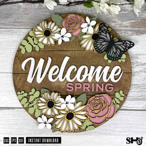 May include: A circular wooden sign with a dark brown plank background featuring the white text Welcome and the pink text SPRING. The sign is decorated with layered cutouts of pink roses, white daisies, green leaves, and a black butterfly.