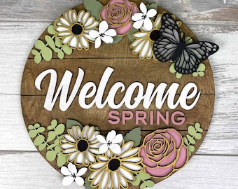 May include: A round wooden sign with the words "Welcome Spring" in white and pink. The sign is decorated with flowers, greenery, and a black butterfly. The sign is a spring-themed decoration.