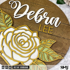 Op de afbeelding: Een houten bord met de naam "Debra Lee" in witte letters op een bruine achtergrond. Een witte roos met gouden contouren en groene bladeren bevindt zich in de linkerbenedenhoek.