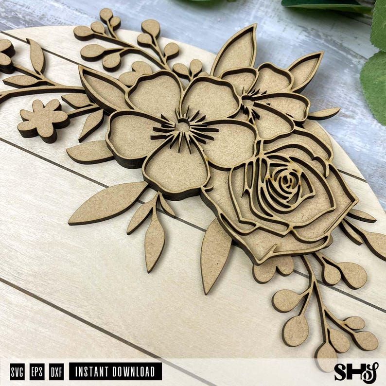 Puede incluir: Un arreglo floral de madera cortada con l&aacute;ser en color marr&oacute;n claro, con p&eacute;talos, hojas y tallos superpuestos. El dise&ntilde;o incluye una flor central con estambres detallados y una flor tipo rosa, montadas sobre una base circular de madera plana.