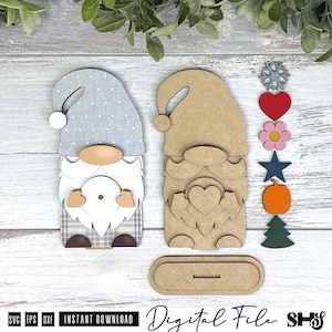 May include: Two gnome figures and six interchangeable icons including a snowflake, heart, flower, star, pumpkin, and tree. One gnome is finished with a grey polka dot hat and plaid base, while the other is unfinished wood with a heart cutout.