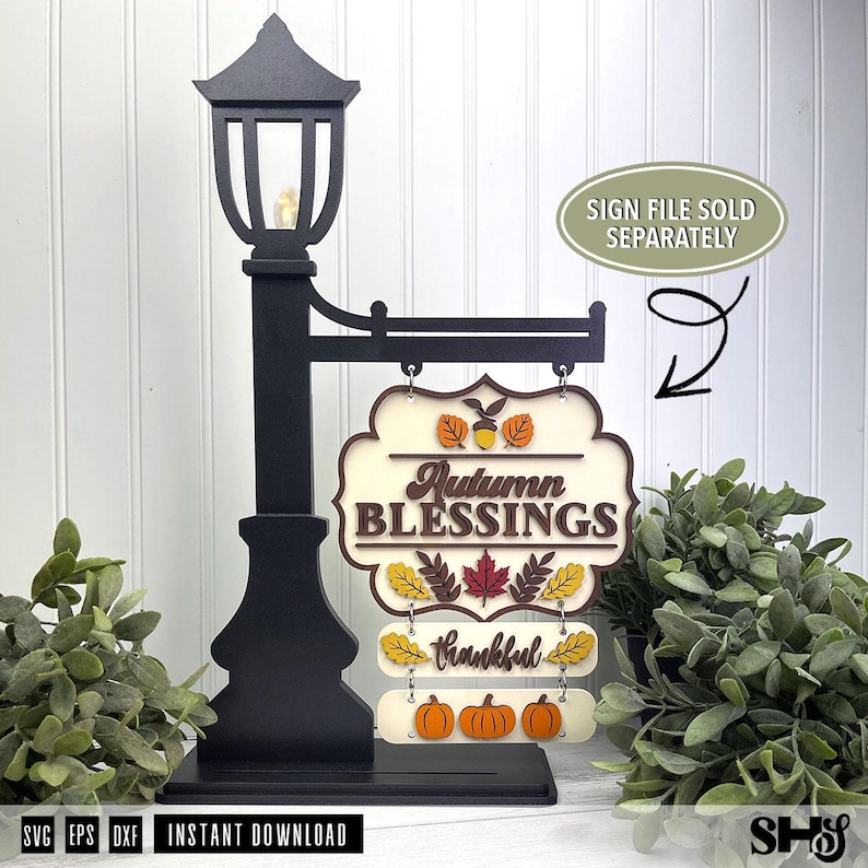 Op de afbeelding: Een zwarte lantaarnpaalstandaard met een hangend bord met de tekst Autumn BLESSINGS en thankful. Het bord is versierd met gelaagde uitsparingen van oranje, gele en rode herfstbladeren, een eikel en drie oranje pompoenen aan de onderkant.