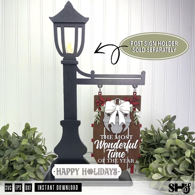 May include: A black decorative lamp post sign holder with a hanging brown wooden sign featuring a white bell, a white bow, and the text THE MOST WONDERFUL TIME OF THE YEAR. A white base plate reads HAPPY HOLIDAYS. Text indicates post sign holder sold separately.