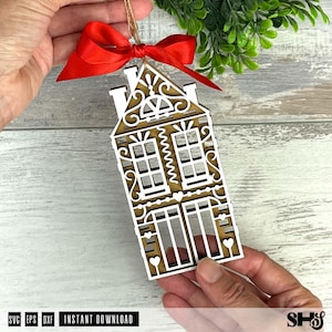 Puede incluir: Adorno de casa de madera cortada con l&aacute;ser con intrincados detalles blancos y un fondo marr&oacute;n. El adorno est&aacute; adornado con una cinta roja y una cuerda para colgar. El texto "SVG EPS DXF INSTANT DOWNLOAD" es visible en la parte inferior.