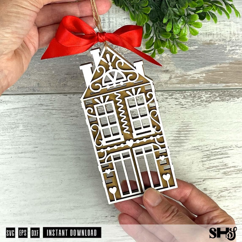 Puede incluir: Adorno de casa de madera cortada con l&aacute;ser con intrincados detalles blancos y un fondo marr&oacute;n. El adorno est&aacute; adornado con una cinta roja y una cuerda para colgar. El texto "SVG EPS DXF INSTANT DOWNLOAD" es visible en la parte inferior.