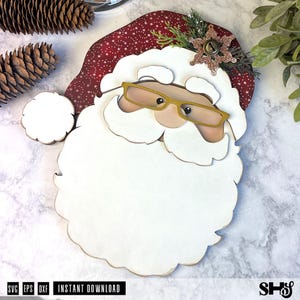 K&ouml;nnte beinhalten: Eine mehrschichtige Holzdekoration in Form eines Weihnachtsmann-Gesichts mit roter M&uuml;tze mit wei&szlig;en Sternen, einem gro&szlig;en wei&szlig;en Bart und einer goldfarbenen Brille. Die M&uuml;tze ist mit einer glitzernden bronzefarbenen Schneeflocke und Tannenzweigen verziert.