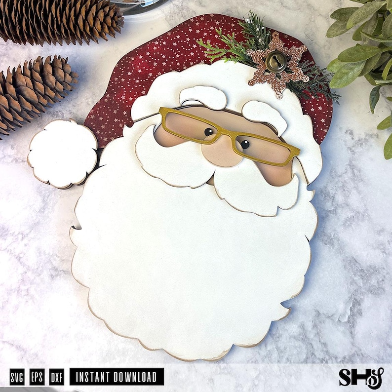 K&ouml;nnte beinhalten: Eine mehrschichtige Holzdekoration in Form eines Weihnachtsmann-Gesichts mit roter M&uuml;tze mit wei&szlig;en Sternen, einem gro&szlig;en wei&szlig;en Bart und einer goldfarbenen Brille. Die M&uuml;tze ist mit einer glitzernden bronzefarbenen Schneeflocke und Tannenzweigen verziert.