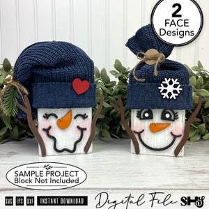 May include: Two wooden block snowman decorations featuring navy blue knit hats. One snowman has a red heart, the other a white snowflake. Both have orange carrot noses, brown twig arms, and painted faces. Text includes 2 FACE DESIGNS and SAMPLE PROJECT.