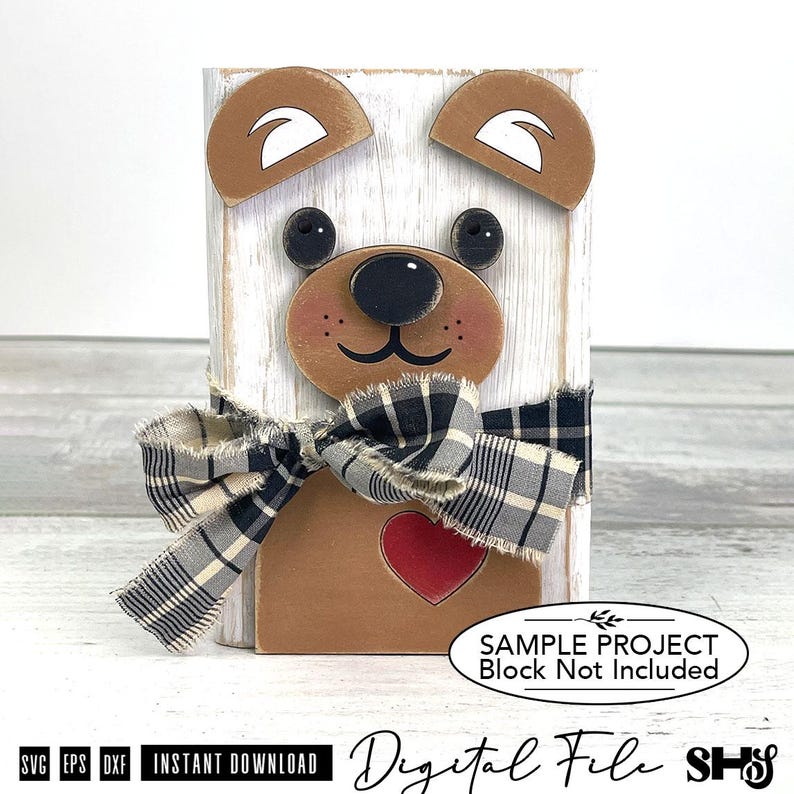May include: A wooden block craft featuring a bear face with brown ears, black eyes, a black nose, and a red heart. A black and white plaid fabric ribbon is tied around the center. The text reads: SAMPLE PROJECT Block Not Included.