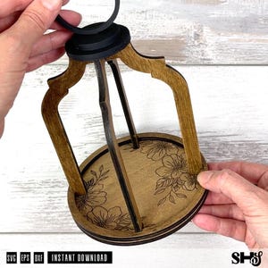 May include: A wooden lantern frame with a circular base featuring laser-engraved floral patterns. The structure consists of curved brown wooden uprights connected to a black top ring. The design includes a flat base with detailed flower illustrations.