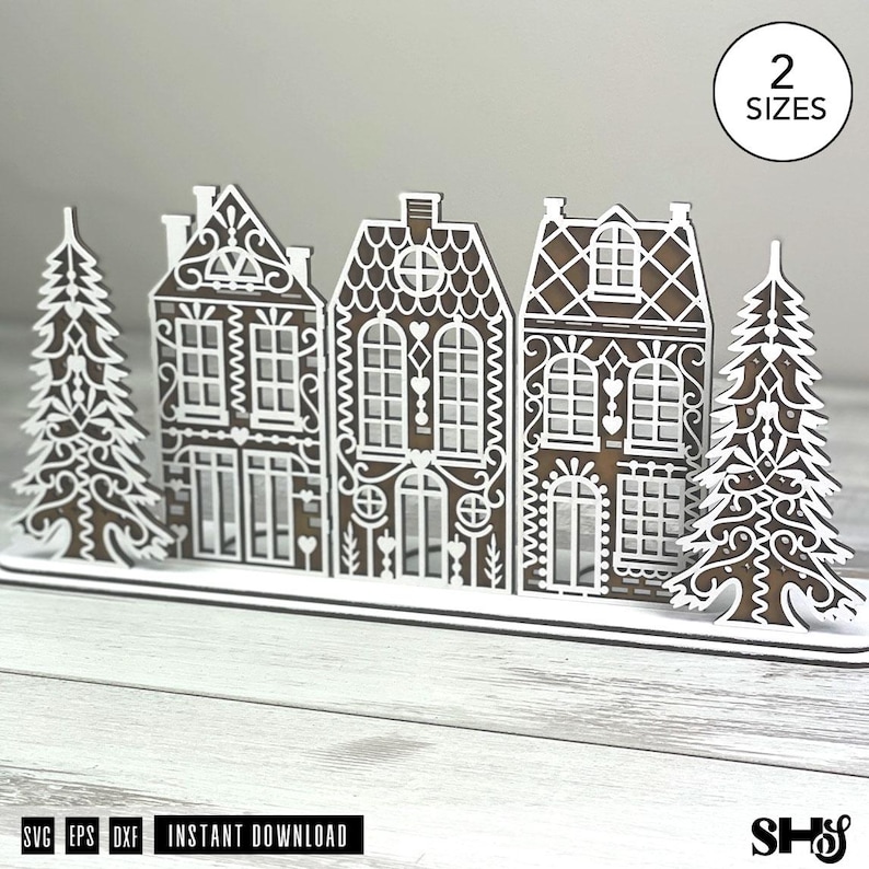 May include: A brown and white laser-cut wooden display featuring three gingerbread-style houses flanked by two evergreen trees. The intricate white filigree patterns mimic icing details on the structures. A circular badge reads 2 SIZES.