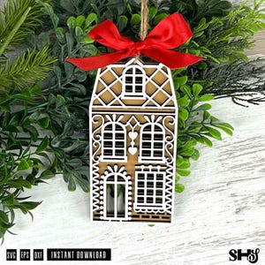 Puede incluir: Adorno de casa de jengibre de madera con intrincados detalles calados blancos y un lazo de cinta roja. El adorno cuelga de una cuerda de hilo natural. El texto "SVG EPS DXF INSTANT DOWNLOAD" es visible.