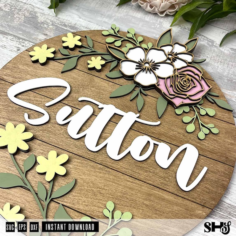 Puede incluir: Un letrero circular de madera con el nombre Sutton en letras cursivas blancas grandes. El letrero est&aacute; decorado con recortes florales de madera en capas en tonos amarillo p&aacute;lido, verde salvia, blanco y rosa suave, dispuestos alrededor del texto.