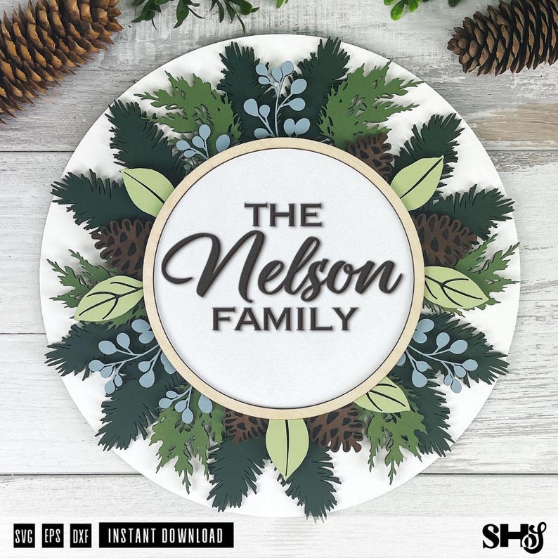 May include: A circular wooden sign featuring a layered design of dark green pine branches, light green leaves, blue berries, and brown pinecones surrounding a central white circle with the black text THE Nelson FAMILY in a mix of fonts.