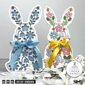 May include: Two white, laser-cut bunny decorations with floral designs and colourful bows. One has a blue bow and blue and white flowers, the other has a yellow bow and multi-coloured flowers. The text on the image says "2 sizes 7-inch & 8.5-inch".