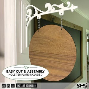Peut inclure: Un panneau circulaire en bois vierge suspendu par deux chaînes en métal argenté à un support mural décoratif blanc. Le panneau présente une finition en grain de bois brun clair lisse. Le texte EASY CUT & ASSEMBLY HOLE TEMPLATE INCLUDED est visible.
