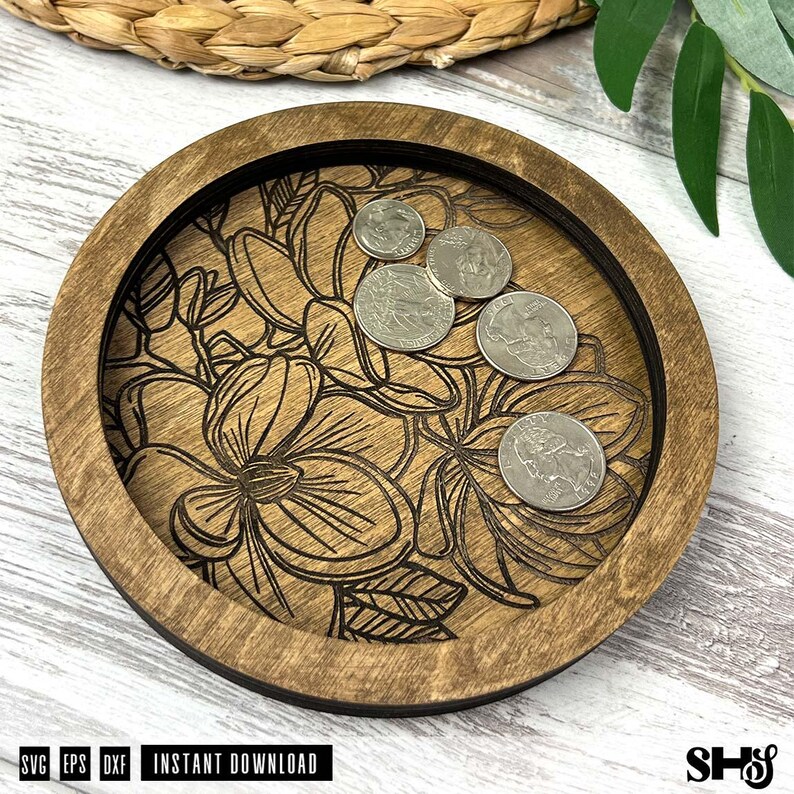 May include: A round wooden tray with a dark brown finish featuring a laser-engraved floral pattern on the base. Four silver-colored coins are resting on the surface of the tray. The tray has a raised circular rim and a flat bottom.