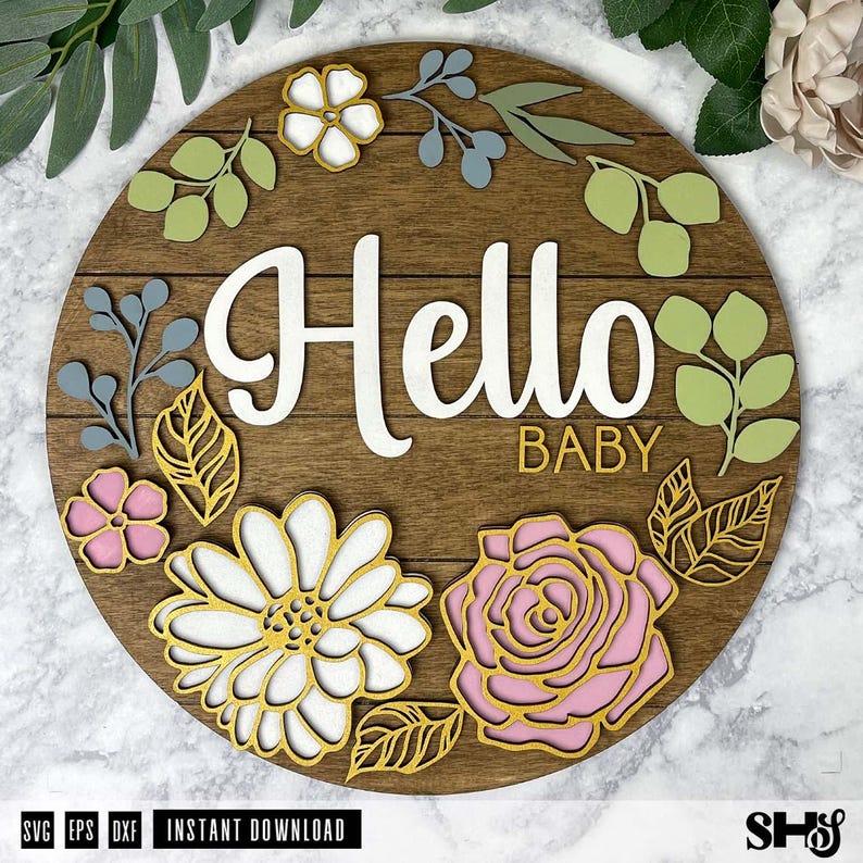 May include: A circular wooden sign featuring the white script text Hello and the gold block text BABY. The sign is decorated with layered wooden cutouts of pink and white flowers, green leaves, and blue sprigs arranged around the lettering.